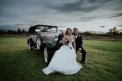 Novios felices en un con nubes de fondo y pasto verde arriba de un auto clasico